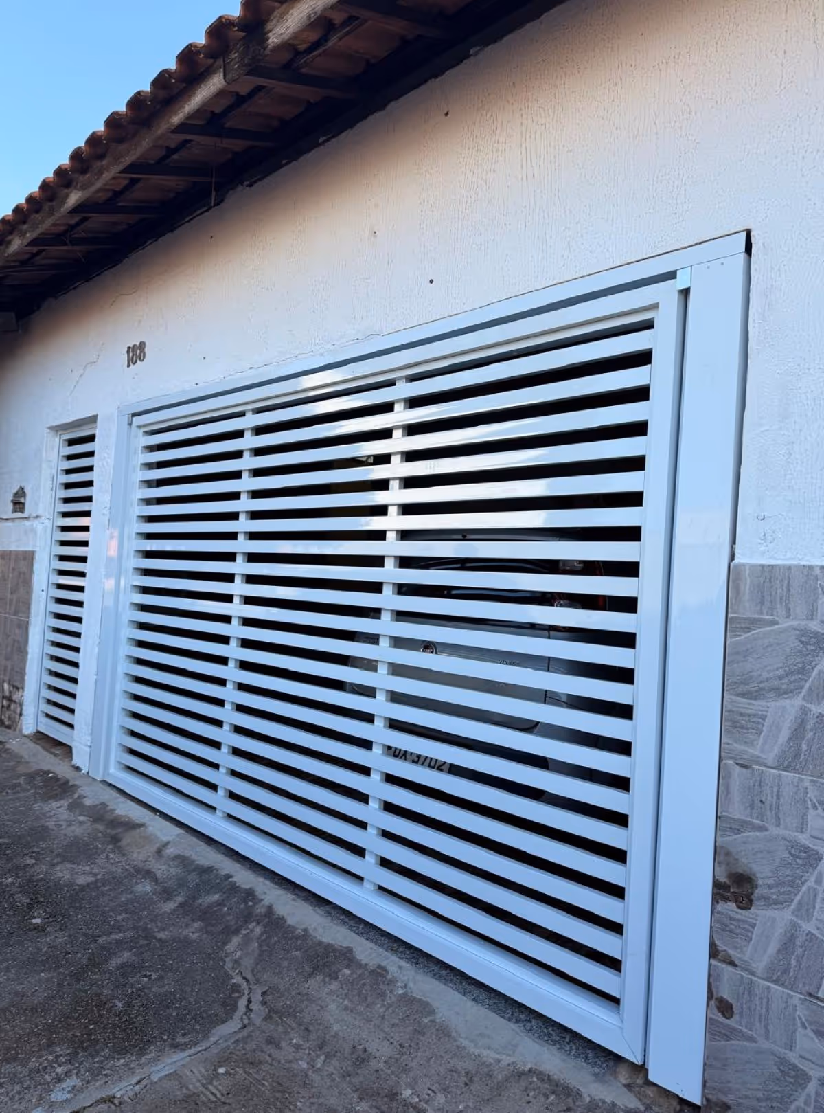 Portão branco de grades horizontais com carro branco estacionado atrás, em frente a uma casa com muro branco e telhado de cerâmica.