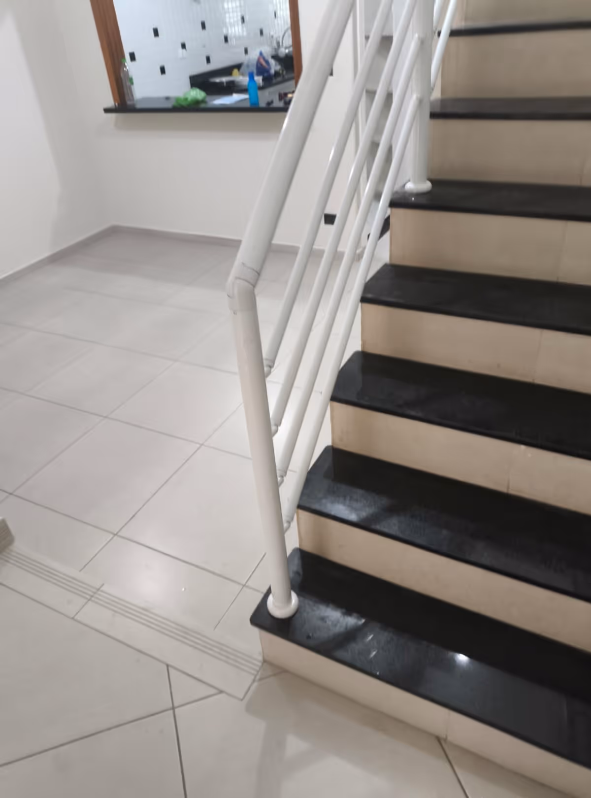 Escada interna com degraus em pedra preta e corrimão branco, próximo a um piso cerâmico claro e uma cozinha ao fundo.