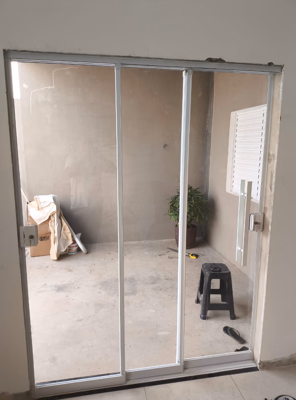 Porta de vidro deslizante branca com três painéis, vista para área externa com parede bege, banquinho preto, planta em vaso, caixa de papelão e chinelo.