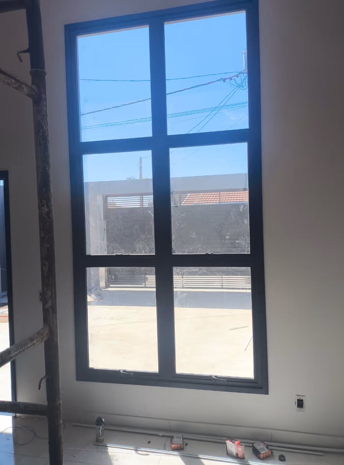 Janela grande de vidro com estrutura preta em uma parede branca, mostrando vista para área externa e céu azul, com materiais de construção no chão.