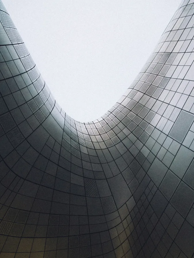 Curved, modern architectural structure with grid-patterned panels viewed from below against a pale sky.